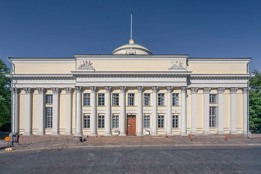 Historic neoclassical building with tall columns and a domed roof, surrounded by trees and a cobblestone walkway. Clear blue sky above.