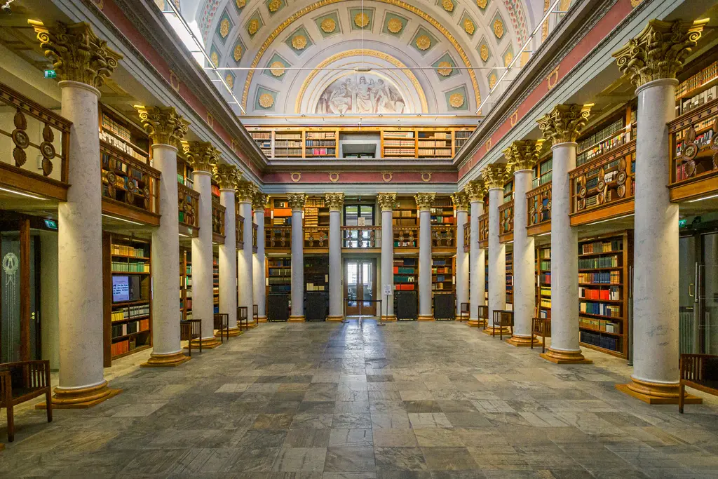 An elegant library interior featuring grand columns, colorful bookshelves, and a beautifully painted ceiling. Cozy reading chairs are arranged throughout.
