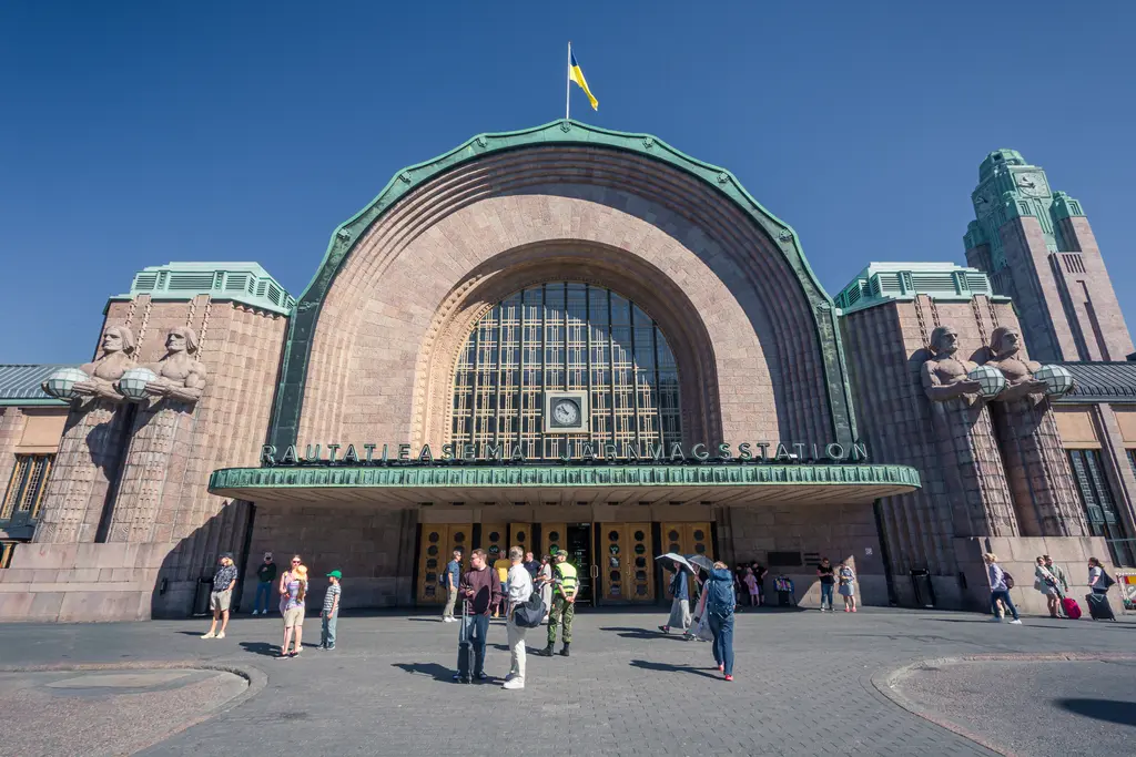 Historic railway station entrance with grand architecture, featuring sculptures and a large arch, bustling with visitors.