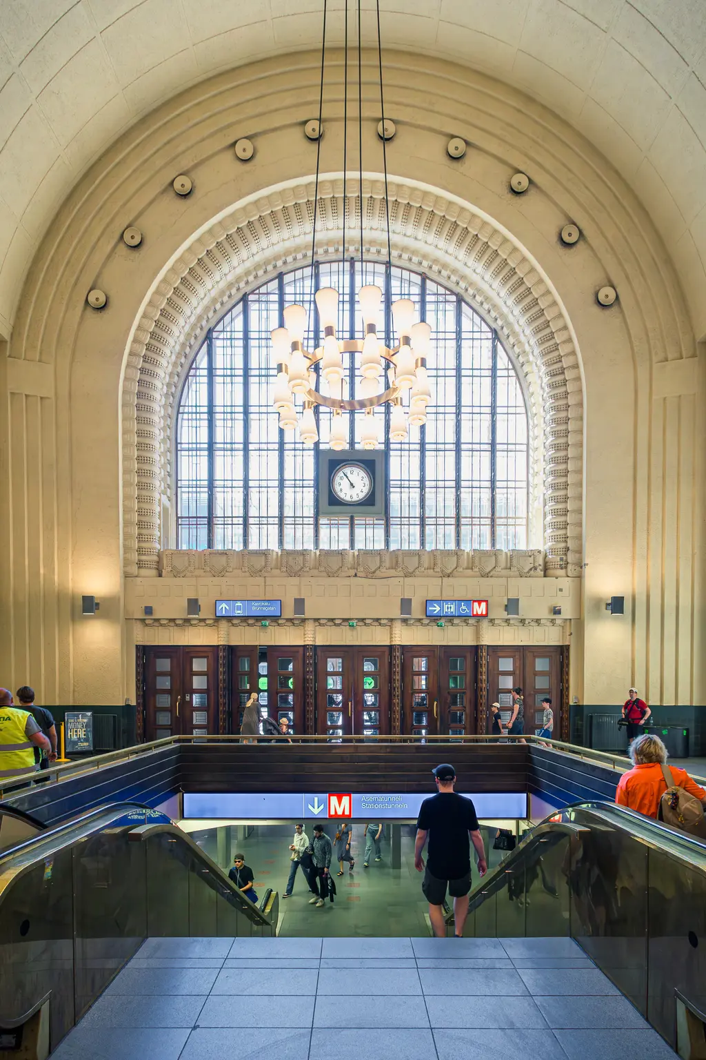 A bustling transit station interior featuring an ornate archway, large clock, and escalators with people moving in various directions.