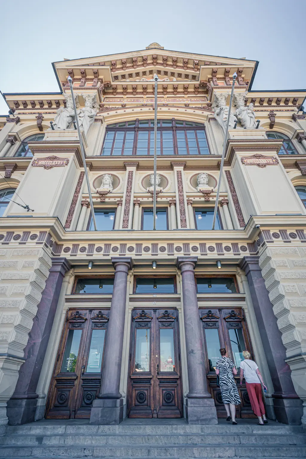 Historic building entrance with ornate wooden doors and classical architecture; two visitors approaching the steps.