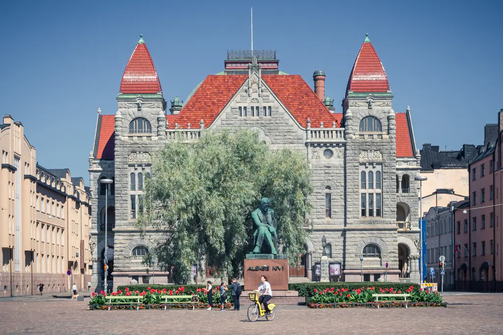 A historic stone building with red roofs and a central statue, surrounded by flowers and people on bicycles in a vibrant urban square.