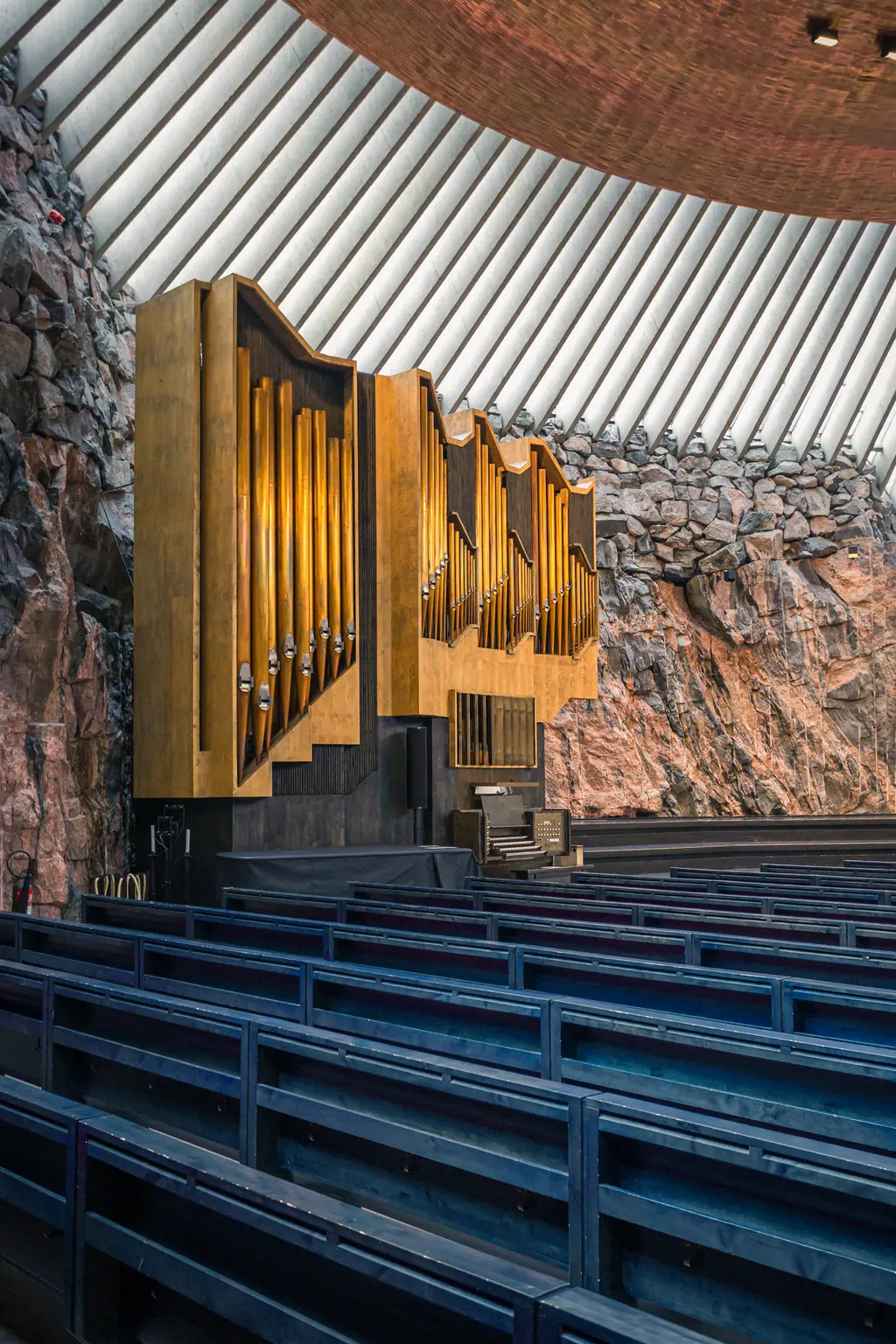 An interior view of a unique rock church featuring a large wooden organ, surrounded by dark stone walls and blue pews.