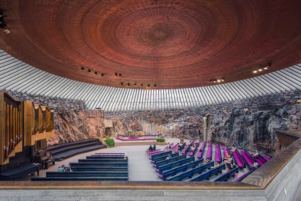 An interior view of a unique concert hall with a wooden dome, rock walls, and purple seating, showcasing modern architectural design.