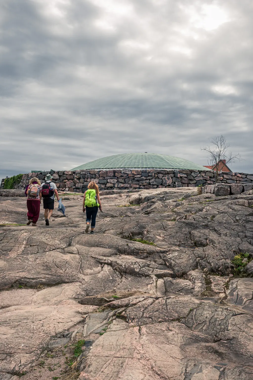 Hikers walking on rocky terrain leading to a unique stone structure under a cloudy sky in Helsinki.