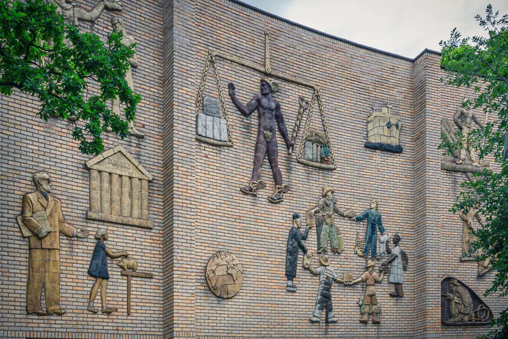 A wall mural featuring various relief sculptures, including figures holding scales and engaged in work, surrounded by greenery.