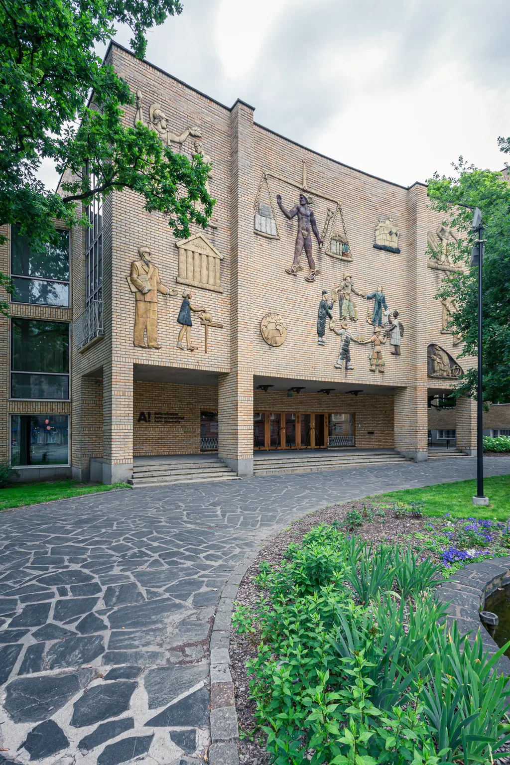 A distinctive brick building featuring artistic reliefs on its facade, surrounded by greenery and a stone pathway.