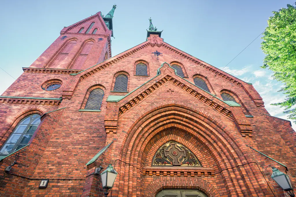 A tall brick church with a prominent steeple and intricate arches, set against a clear sky and surrounded by lush greenery.