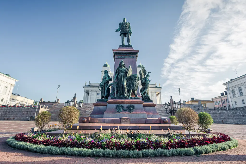 A bronze statue surrounded by colorful flowers, depicting historical figures in a public square with historic buildings in the background.