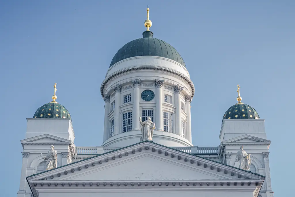 A grand white cathedral roof with a green dome and golden accents under a clear blue sky. Statues adorn the front façade.