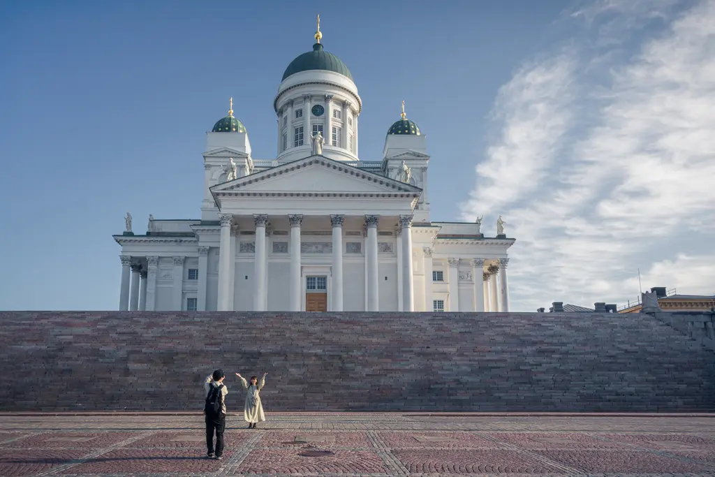 A large white neoclassical building with green domes and a staircase, featuring two people in front, enjoying the view.