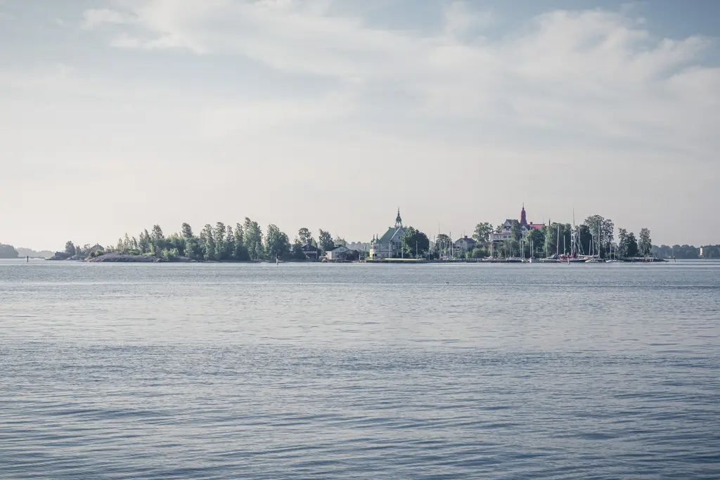 A tranquil view of an island with lush trees and historic buildings, set against calm waters and a soft sky.
