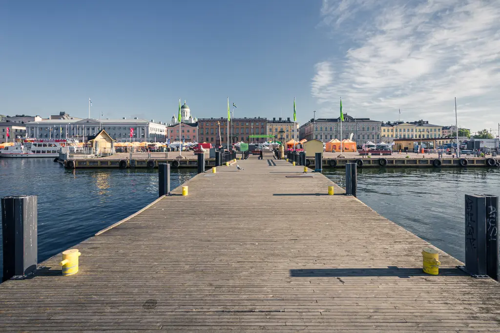 A wooden pier extending into a calm harbor, surrounded by colorful buildings and boats under a clear blue sky.