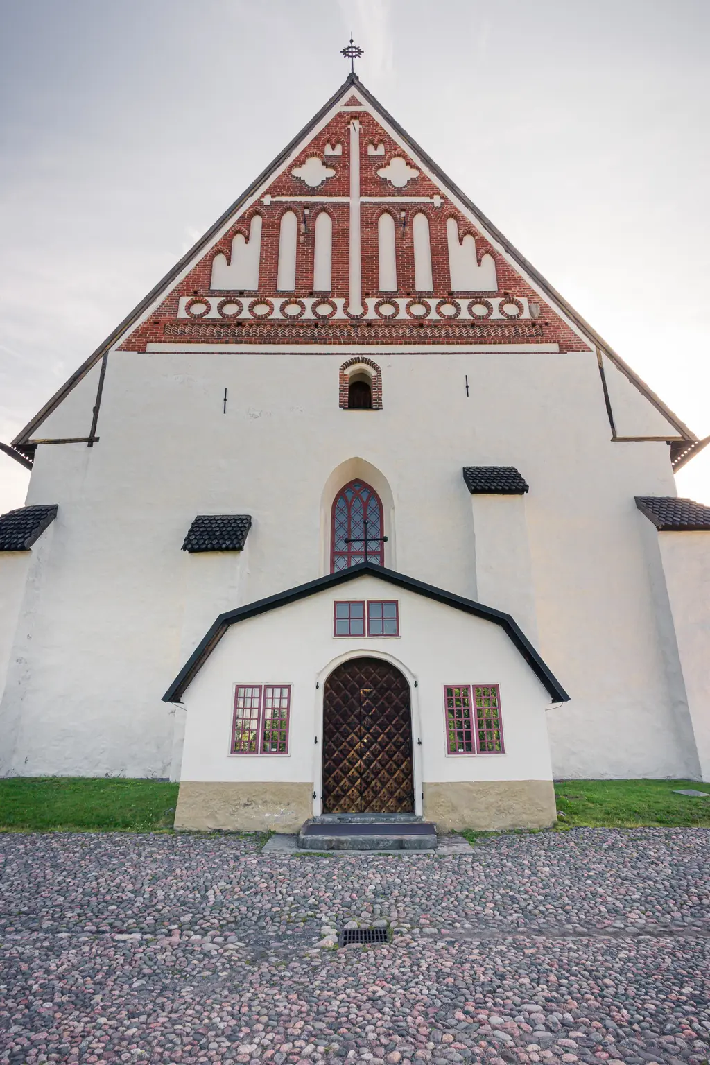 Fassade einer historischen Kirche mit einem weiß gestrichenen Unterbau und einem dreieckigen, gemusterten Dach. Eingangsbereich mit Holzporte.
