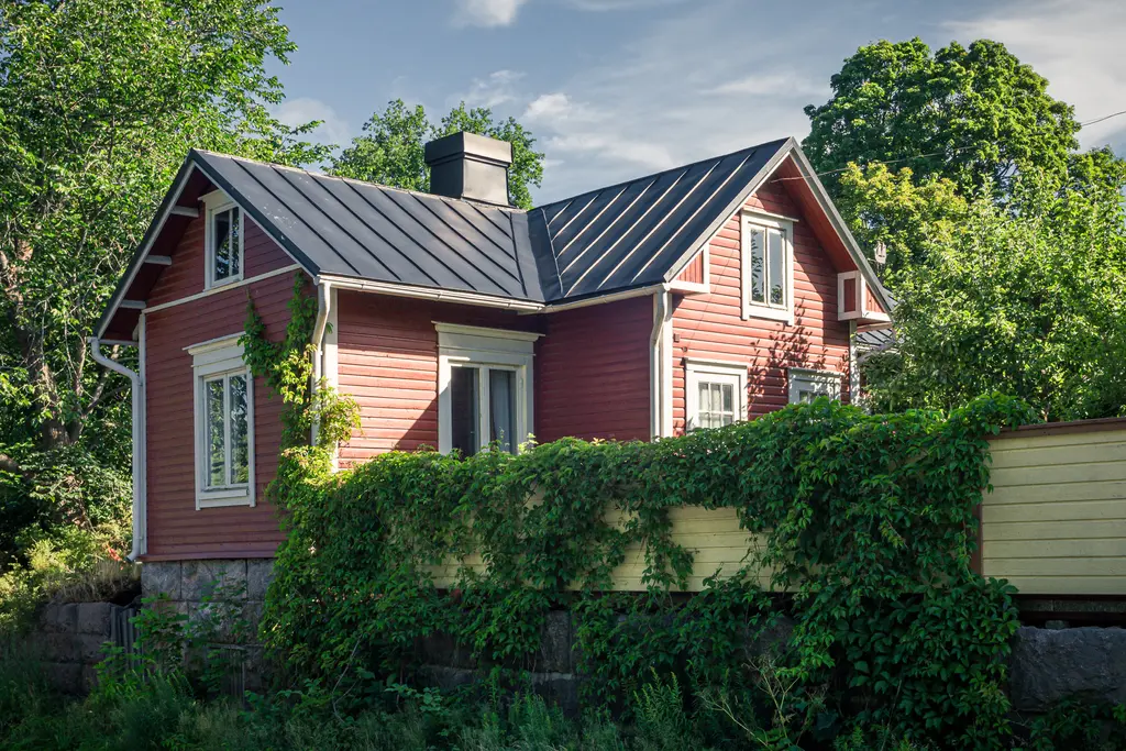 Rotes Holzhaus mit einem schwarzen Dach, umgeben von üppigem Grün und Efeu. Große Fenster lassen Licht hinein.