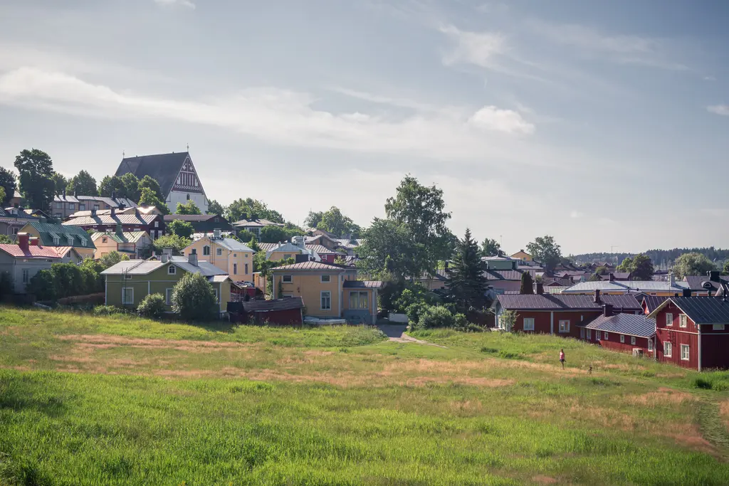 Blick auf eine malerische, hügelige Stadtlandschaft mit bunten, historischen Häusern und einer Kirche im Hintergrund. Grüne Wiesen umgeben die Gebäude.