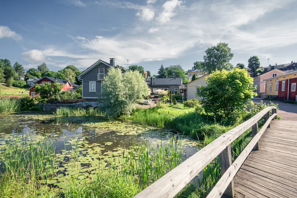 Der Blick über eine kleine Brücke führt zu einem ruhigen Teich, umgeben von üppigem Grün und bunten Häusern im Hintergrund.