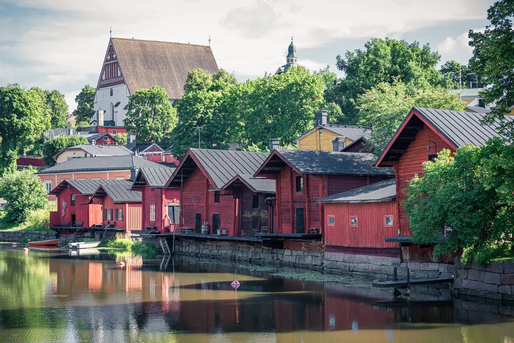Rote Holzhäuser am Ufer eines ruhigen Wassers, umgeben von grünen Bäumen. Im Hintergrund ist eine Kirche mit einem charakteristischen Dach zu sehen.