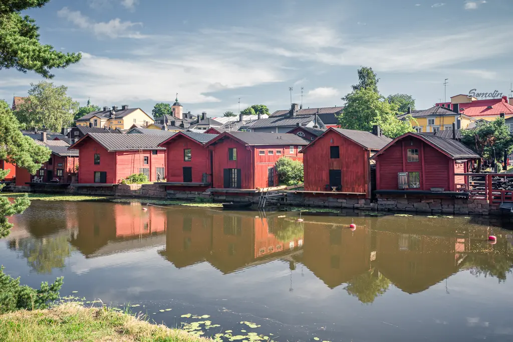 Rote Holzgebäude am Ufer eines ruhigen Gewässers, mit spiegelndem Wasser und grünem Uferbewuchs, umgeben von Bäumen und Häusern.