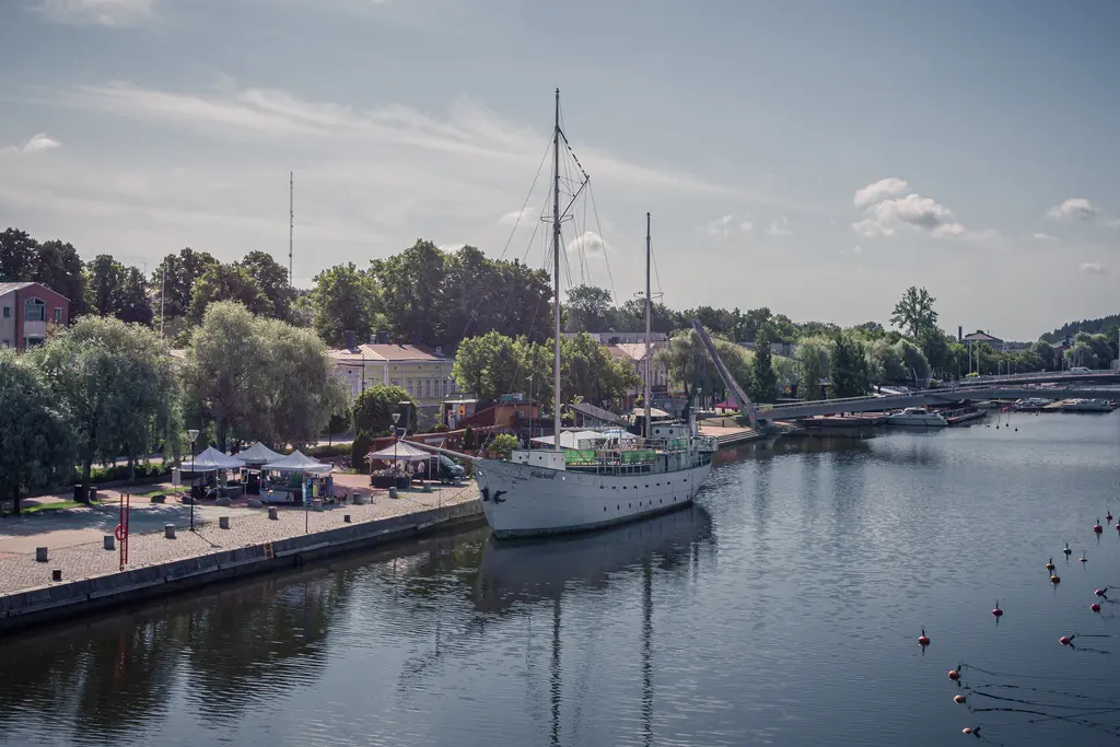 Historisches Segelschiff im ruhigen Wasser, umgeben von Bäumen und einer Uferpromenade mit Ständen und Booten.