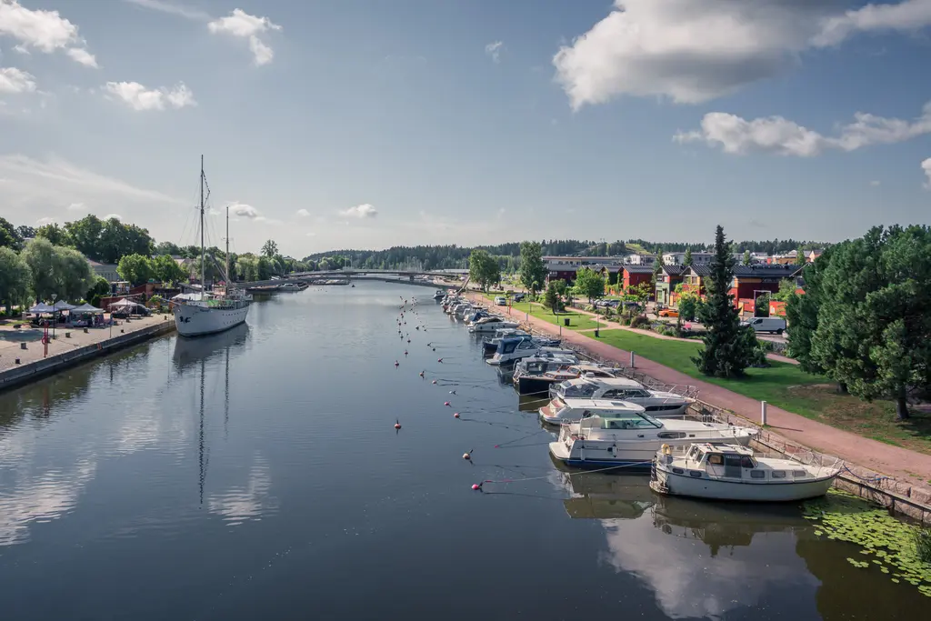 Schöne Hafenansicht mit einem ruhigen Fluss, Booten am Ufer und grünen Bäumen. Helle Wolken spiegeln sich im Wasser.