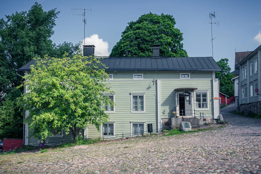 Historisches Holzhaus mit grünlicher Fassade, umgeben von Bäumen. Auf einer gepflasterten Straße sind Antennen auf dem Dach sichtbar.