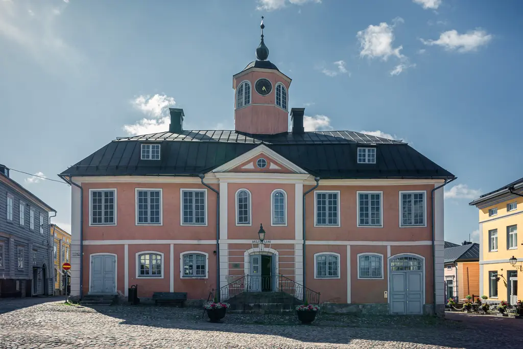 Historisches Gebäude mit bunt gestrichener Fassade, zentralem Uhrturm und Fenster mit weißen Rahmen. Pflasterstraße und Blumenkübel im Vordergrund.