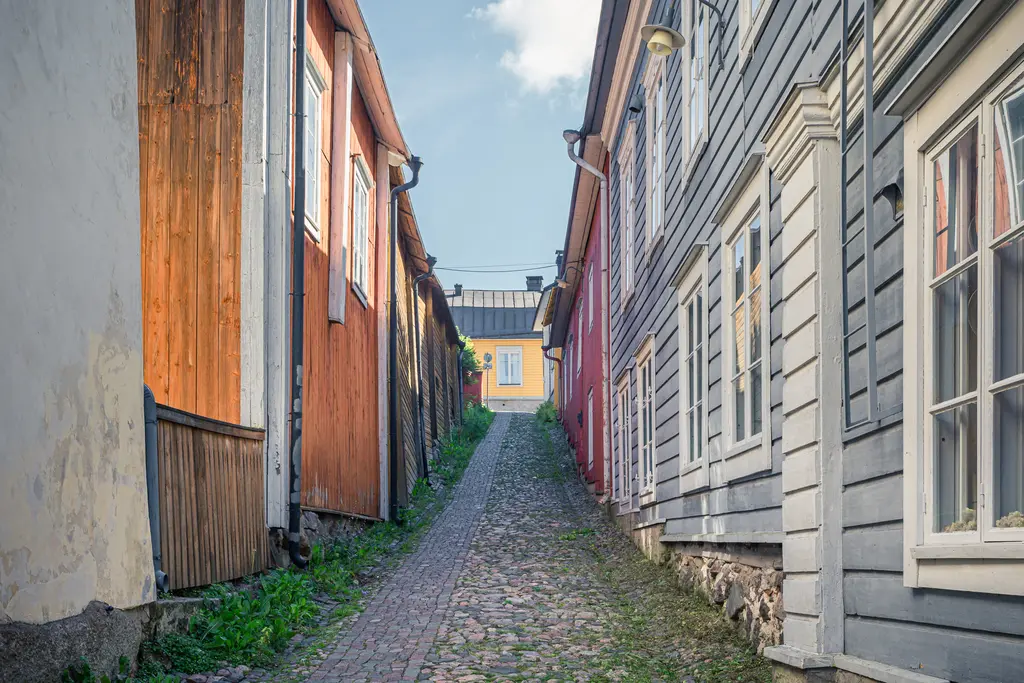 Schmale, gepflasterte Gasse zwischen bunten Holzgebäuden, die sanft ansteigt. Die Wände zeigen orange, grau und gelbe Farben.