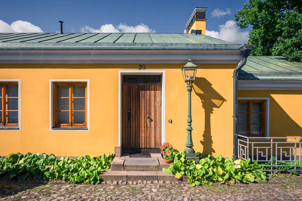 Gelbes Wohnhaus mit grünem Dach, holzverkleideter Eingangstür und Laterne. Umgeben von bepflanzten Beeten und Sichtsteinen.