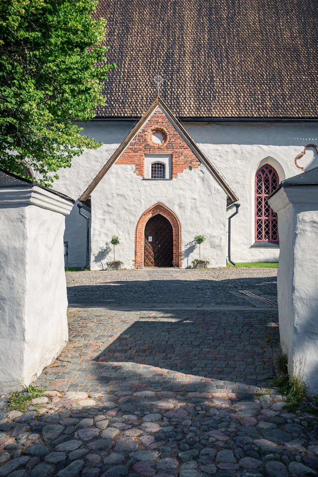 Historische Kirche mit einem großen, holzgetäfelten Eingang und roten Fensterrahmen, umgeben von einem gepflasterten Vorplatz.