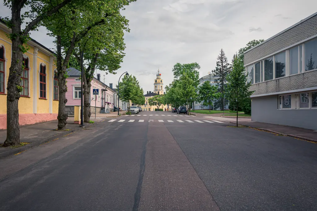 Leere Straße mit Bäumen beidseitig, im Hintergrund ein Turm mit einer markanten Kuppel. Freundliche Atmosphäre in der Stadt.