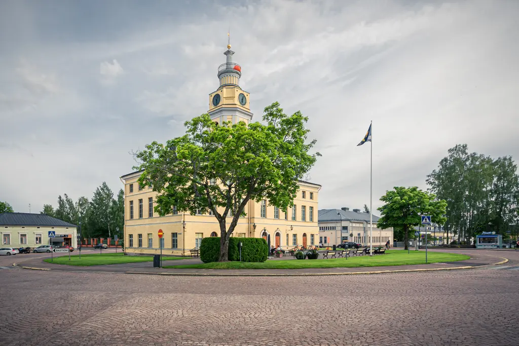 Historisches Gebäude mit Turm und Uhr vor einem großen Baum, umgeben von einer runden Verkehrsinsel und gepflasterter Straße.