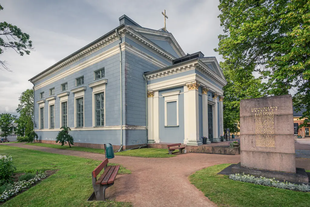 Blick auf eine historische Kirche mit blauer Fassade und weißen Säulen, umgeben von einem gepflegten Garten und Bänken.
