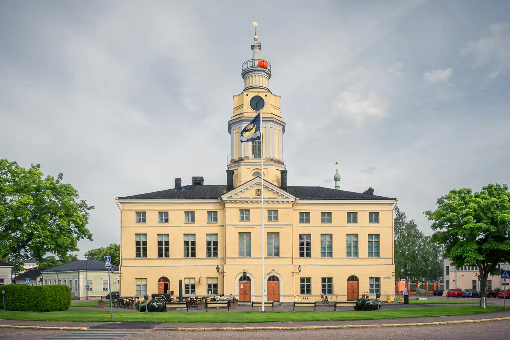 Historisches Gebäude mit gelber Fassade und Uhrturm, umgeben von Bäumen und einem kleinen Platz vor der Eingangstür.