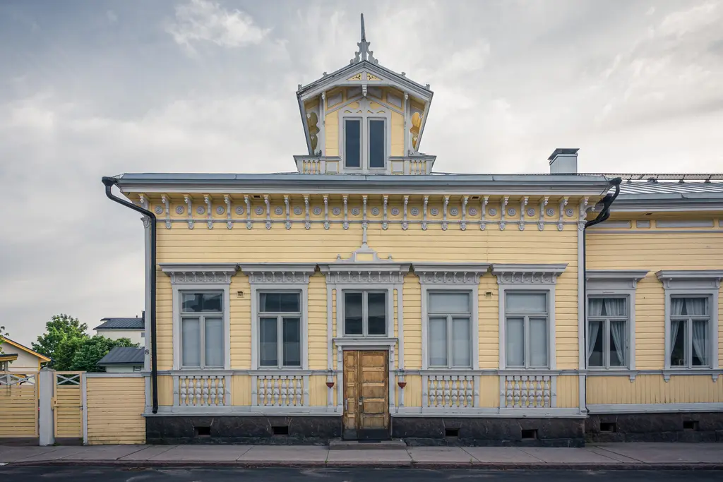 Historisches Holzgebäude mit gelber Fassade, verziertem Dach und mehreren Fenstern. Ein Haupteingang aus Holz ist zentral platziert.