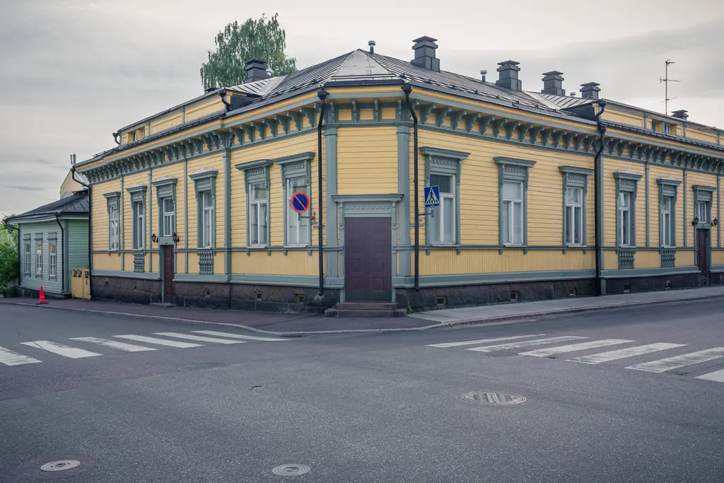 Eckhaus mit gelber Fassade und grünen Verzierungen, mehrere Fenster und ein zentraler Eingang. Nahe einer Straßenecke.