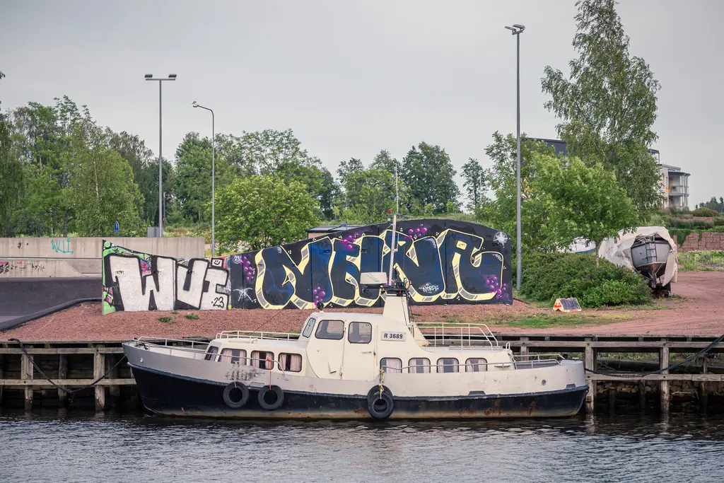 Alter Schiff im Hafen von Hamina, vor einer bunten Graffiti-Wand. Begrünte Ufervegetation und ein ruhiges Gewässer im Hintergrund.