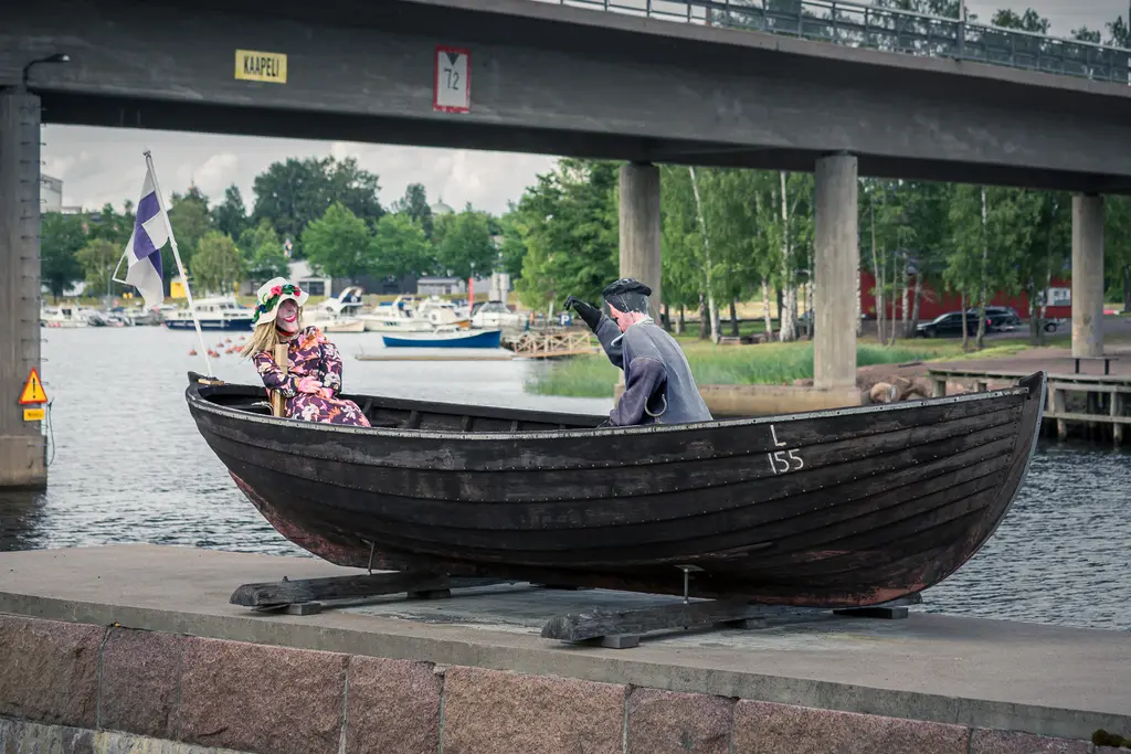 Zwei Holzfiguren in einem kleinen Boot stehen an einem Ufer. Eine Figur trägt bunte Kleidung, die andere ist schlicht gekleidet. Im Hintergrund sind Boote und Bäume sichtbar.