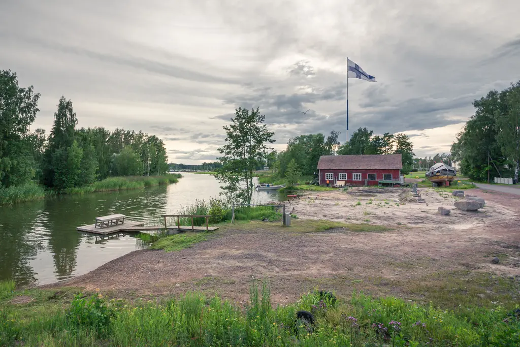 Ruhiger Fluss in Hamina mit einem roten Holzhaus, umgeben von Bäumen. Eine Flagge weht vor dem Gebäude, das Ufer ist sandig.