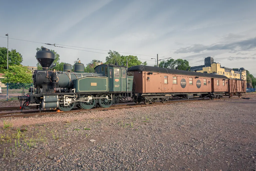 Dampflok mit historischer Lokomotive und Waggon auf Schienen, umgeben von Bäumen und einem modernen Gebäude im Hintergrund.