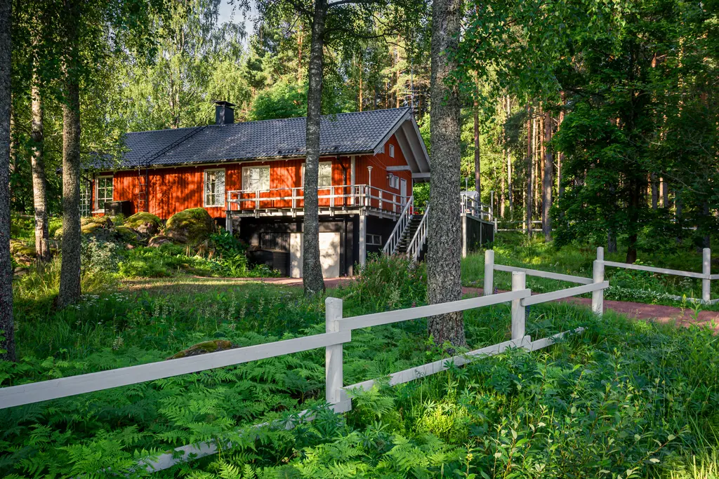 A charming red house surrounded by lush greenery in Virolahti, Finland, with a white wooden fence and natural landscaping.