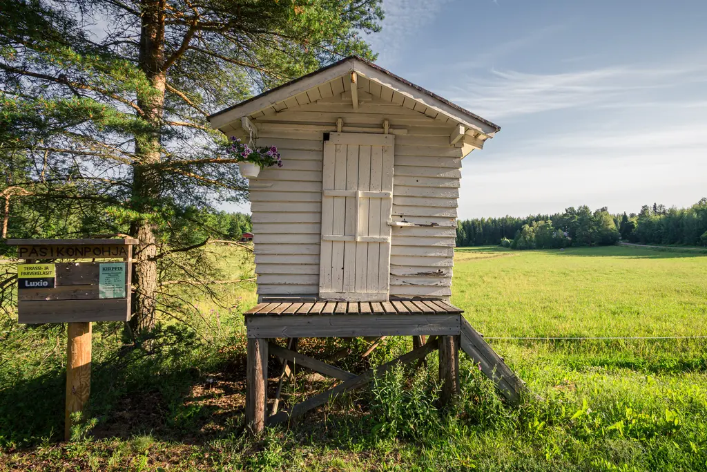 A small wooden hut on stilts in Virolahti, surrounded by greenery and a grassy field under a clear sky.