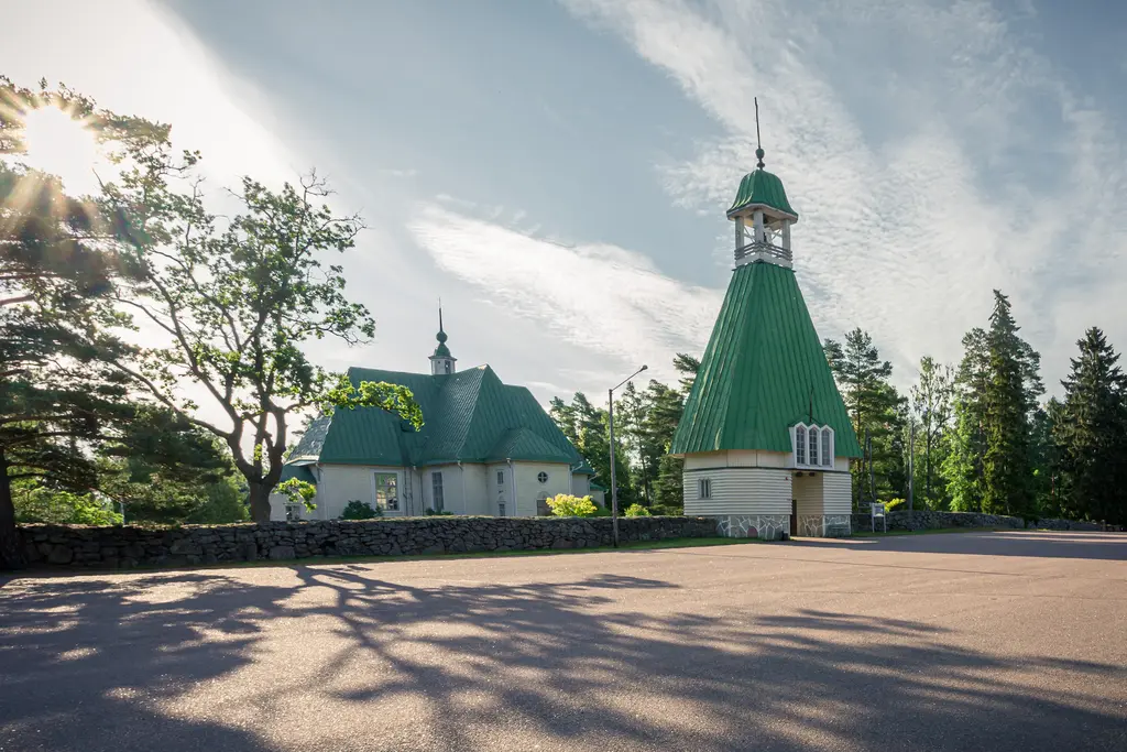 A green-roofed church building in Virolahti surrounded by trees and sunlight, with a bell tower prominently featured.