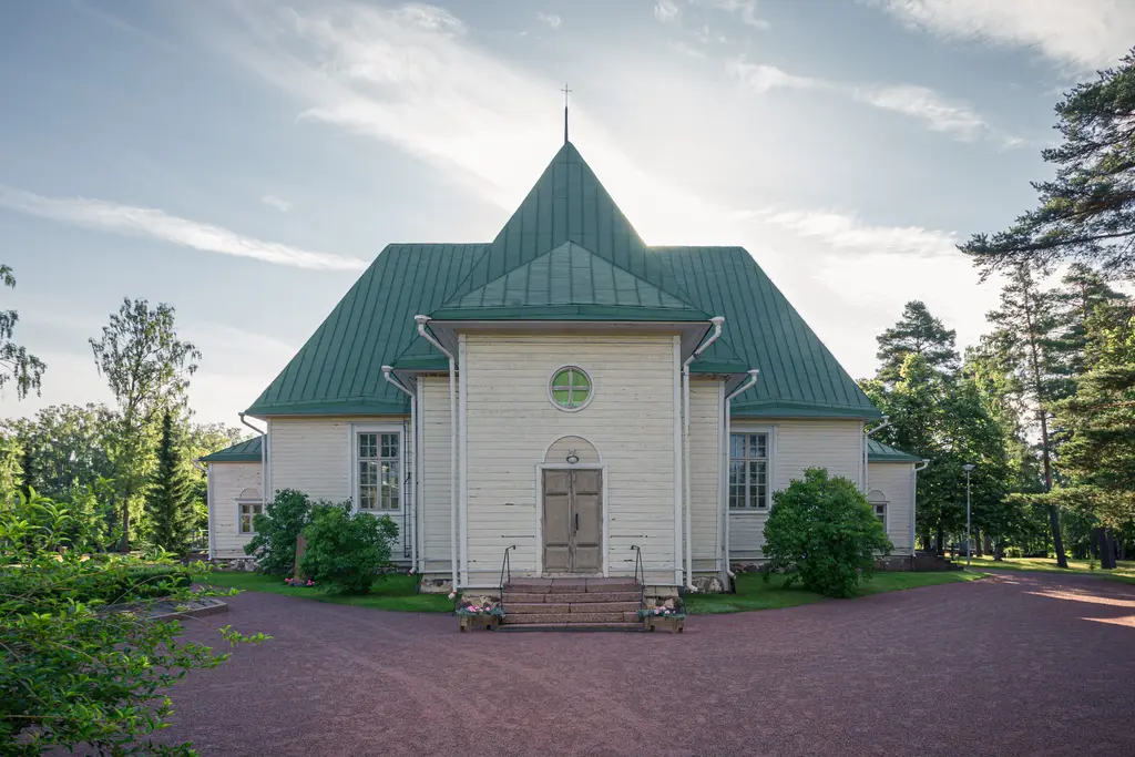A wooden church in Virolahti, Finland, featuring a green triangular roof and surrounded by trees and greenery.