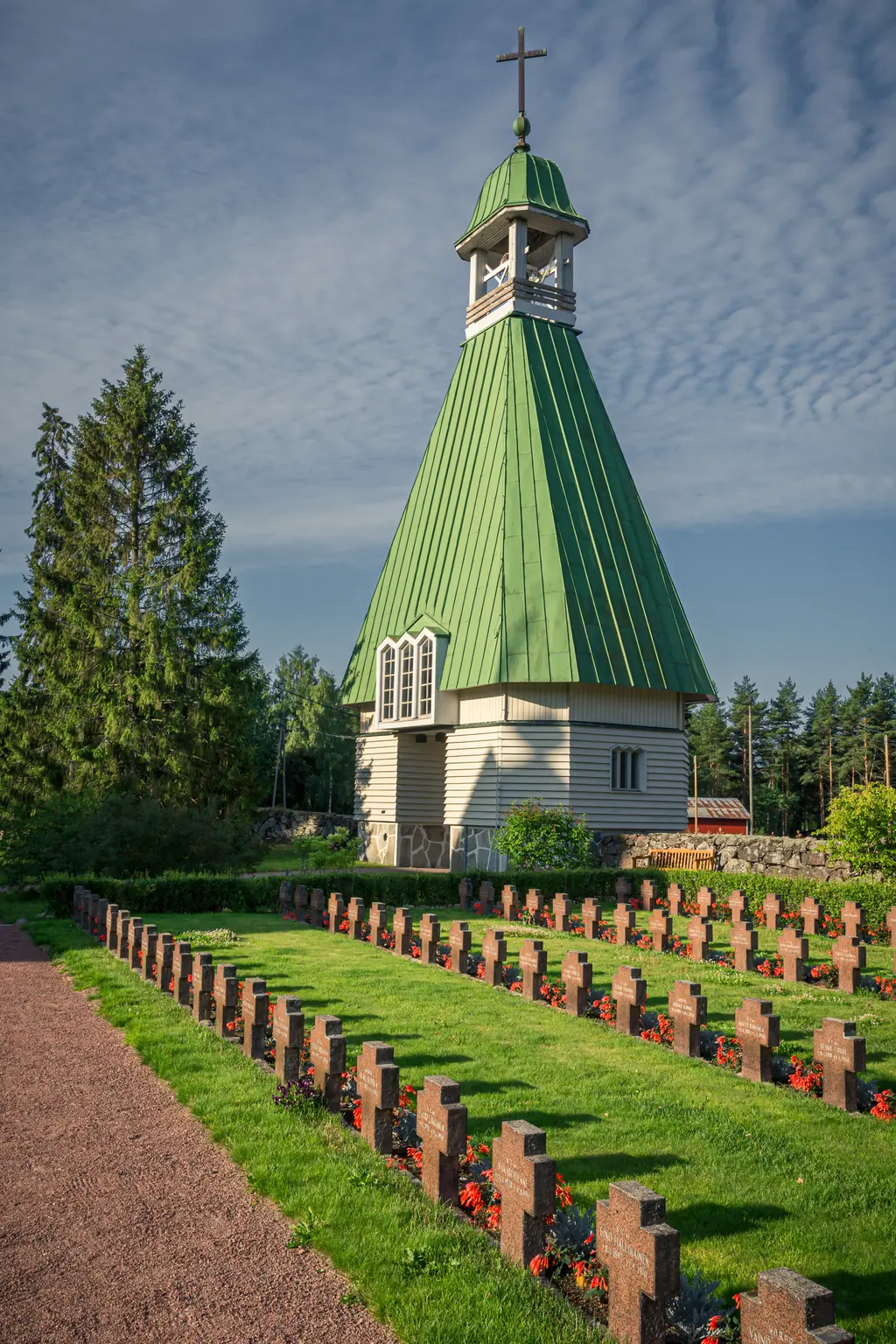 A wooden church with a green steeple in Virolahti, surrounded by a well-maintained cemetery with floral decorations.