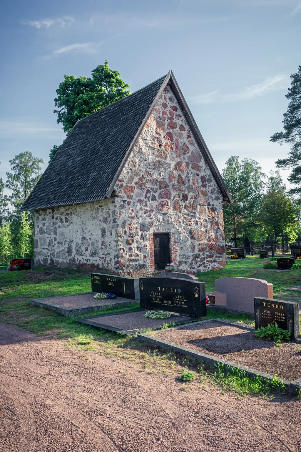 A small stone chapel in Virolahti surrounded by gravestones and greenery, set under a clear blue sky.