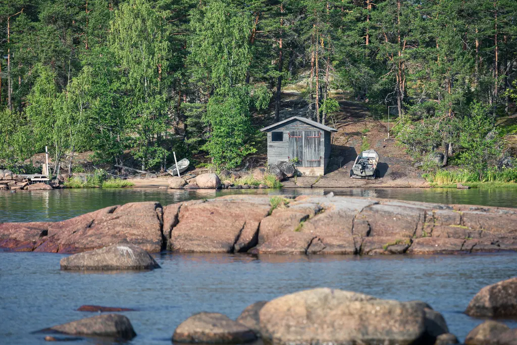 A serene view of rocky shoreline at Virolahti, Baltic Sea, featuring a wooden shed and two boats. Lush greenery surrounds the area.
