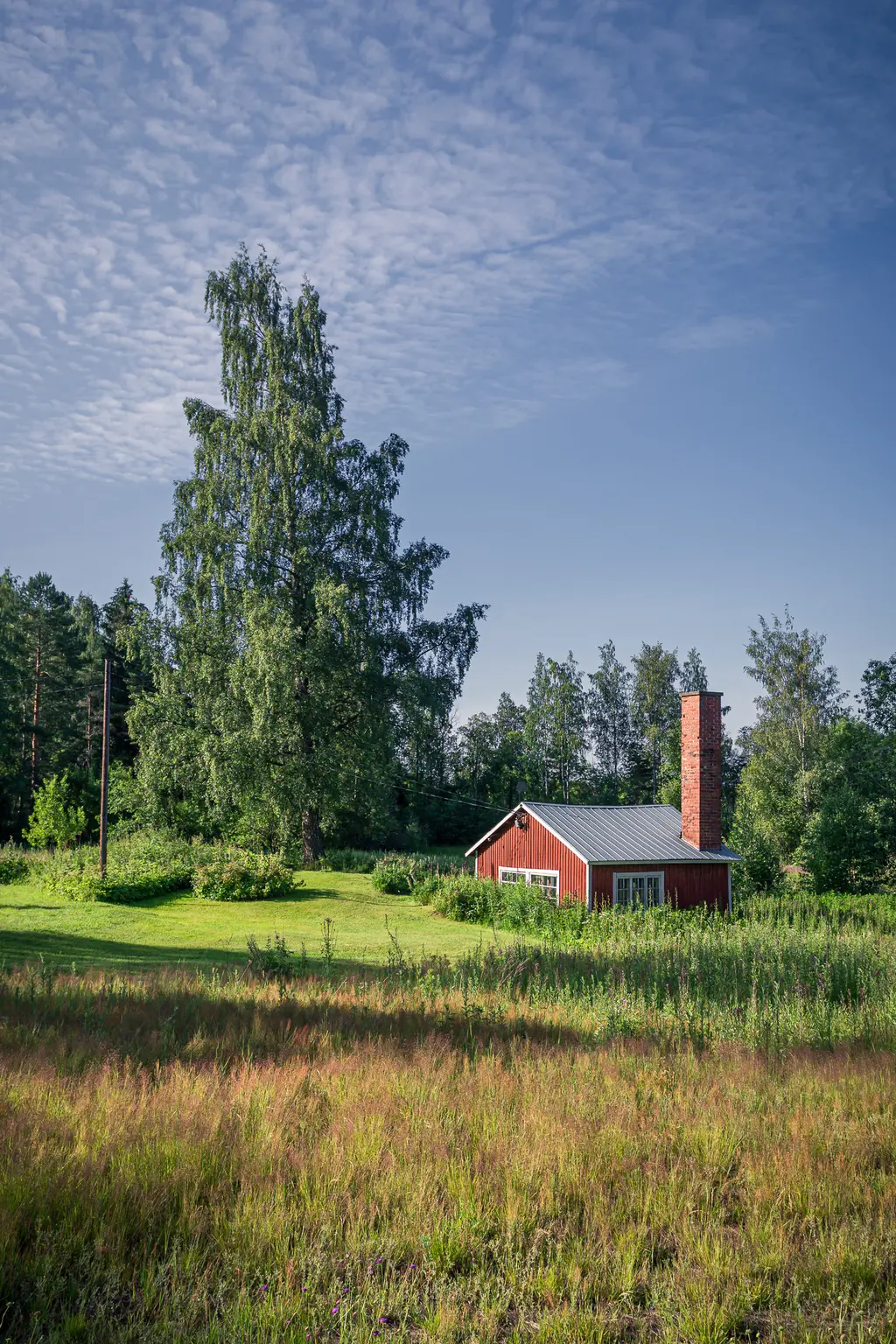 A red cottage among lush green fields in Virolahti, surrounded by tall trees under a clear blue sky.