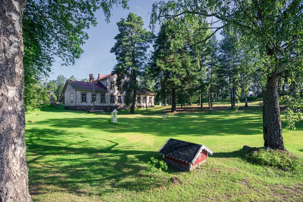 A historic house in Virolahti surrounded by lush green grass and tall trees, with a small red garden shed in the foreground.