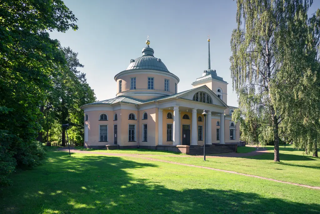 Rundes, helles Gebäude mit einer Kuppel und Säulen, umgeben von Rasenflächen und Bäumen. Die Fassade ist in Pastellfarben gehalten.